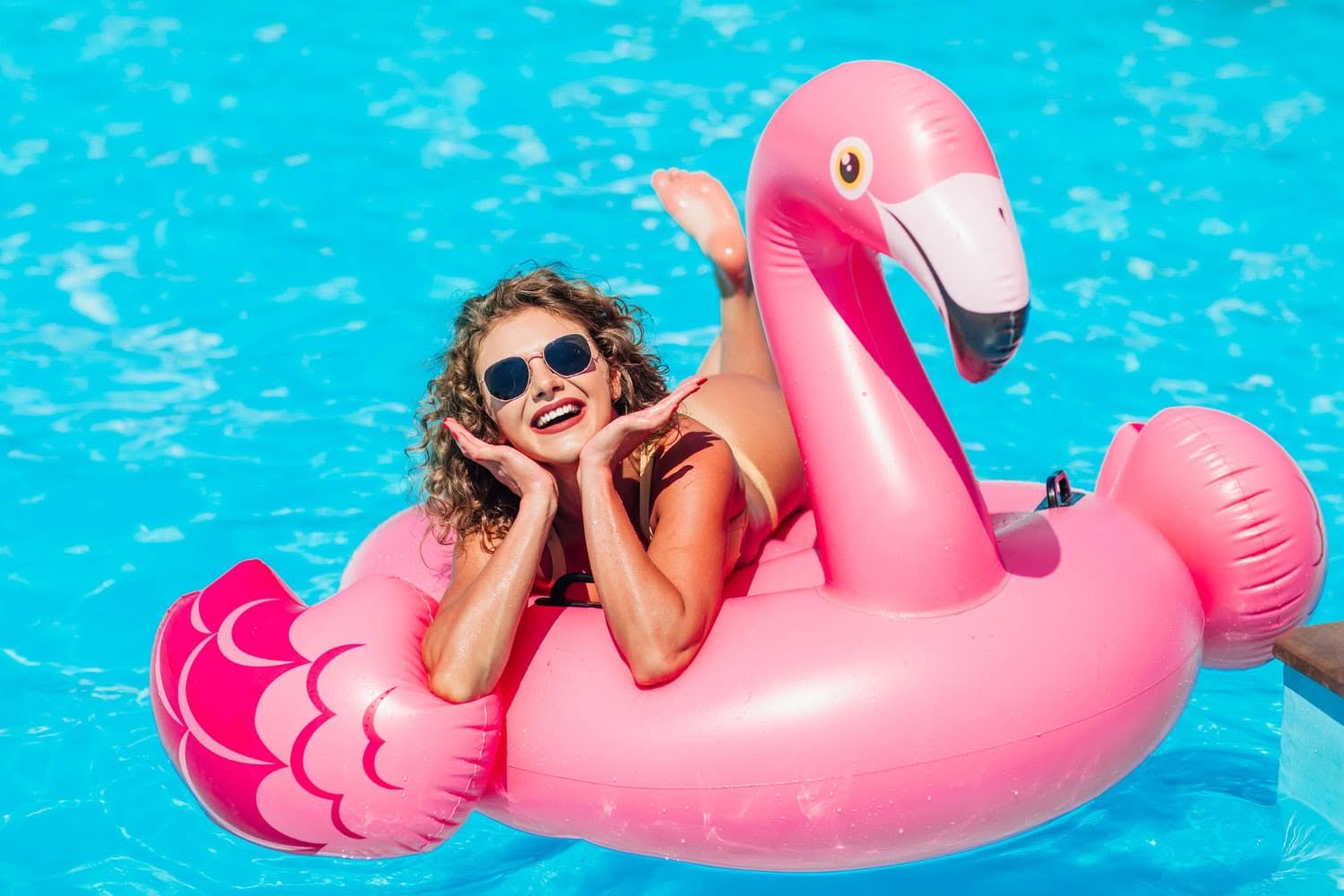 A woman in sunglasses and a red swimsuit smiles while lying on a pink flamingo pool float in a swimming pool, enjoying the sun after following late summer pool care tips from pool experts.