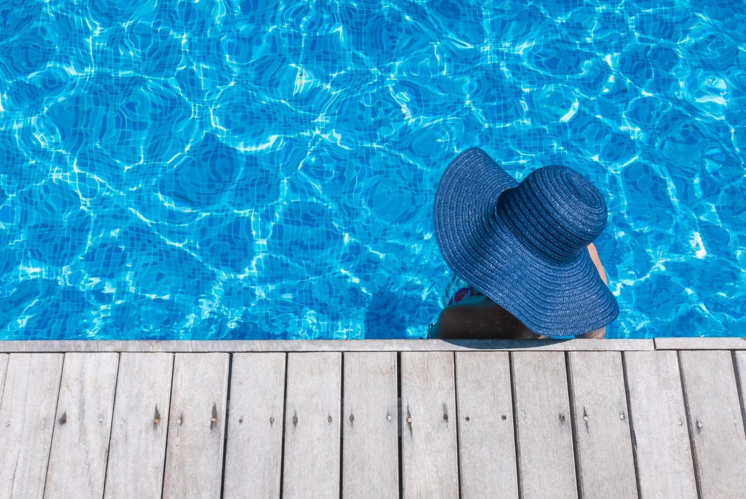 Person wearing a large blue sunhat sits at the edge of a wooden deck, with feet in a clear blue swimming pool, enjoying the peace that comes with reliable pool service contracts.